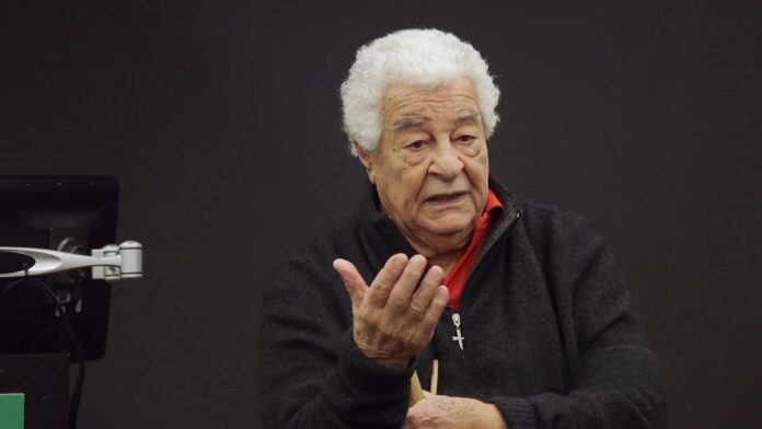Antonio Carluccio speaking during a public discussion, gesturing with one hand while seated against a dark background