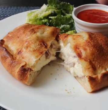 Golden-brown Italian panzerotti cut open to reveal melted cheese and ham, served with marinara dipping sauce and fresh lettuce on a white plate