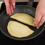 Two uncooked panzerotti cooking in a nonstick skillet on a stovetop, ready to develop a golden, crispy crust
