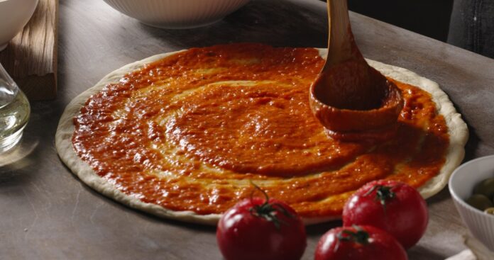 Pizza dough being spread with traditional pizza sauce before toppings are added